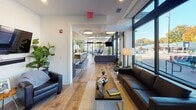 Located at 2604 Hillsborough Ave in Raleigh, NC, this thoughtfully designed single-floor space blends professional functionality with residential comfort. Floor-to-ceiling windows along the street-facing façade flood the interior with natural light and offer open views of the surrounding Hillsborough Avenue corridor, including colorful street murals and mature trees. The open lounge area serves as a welcoming reception and gathering space, featuring warm hardwood-style flooring, leather seating, and a relaxed layout that transitions seamlessly into a secondary collaborative work zone outfitted with pendant lighting and additional seating arrangements. The fully equipped kitchen includes dark espresso cabinetry, stainless steel appliances—including a refrigerator, electric range, and microwave—and a generous white quartz peninsula with bar seating, ideal for hosting or casual breakroom use. A private bedroom provides a quieter retreat, highlighted by a white-painted exposed brick accent wall, gold-toned lighting fixtures, and tasteful artwork that lend a warm, boutique atmosphere. The restroom features a dark vanity with clean white walls and a distinctive patterned tile floor, adding a stylish touch. Throughout the space, crisp white walls, wood-look flooring, and modern finishes create a cohesive, contemporary aesthetic well-suited for a dynamic professional environment.
