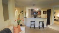 This single-floor residence features an open-concept design that integrates the kitchen and living area into one cohesive space. The kitchen is outfitted with rich dark wood cabinetry, black appliances including a built-in microwave and range, and a decorative tile backsplash. Track lighting illuminates the kitchen, highlighting the dark granite-style countertops that extend to form a raised breakfast bar — a practical and stylish divider between the kitchen and living space. Two black bar stools sit at the bar, offering casual seating. The living area features neutral carpet flooring and warm beige walls that create a welcoming ambiance throughout. Decorative touches, including wall art and a vase of orange tulips, add warmth and personality to the space. A hallway visible from the living area suggests additional rooms beyond. The overall design balances functionality and comfort, with an efficient, well-organized layout suited for everyday living and entertaining.