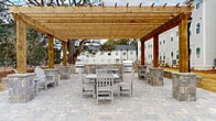 Located at 144 E 62nd St in Savannah, Georgia, this single-story property offers a thoughtfully designed outdoor environment that serves as a central gathering area. The standout feature is a generously sized wood pergola constructed from warm-toned cedar lumber, supported by sturdy posts and topped with an open lattice roof that allows natural light to filter through while providing partial shade. Beneath the pergola, a broad paver patio finished in neutral gray stone tones accommodates multiple seating arrangements, including dining tables with slatted chairs suited for group gatherings. Flanking the seating areas are built-in masonry structures housing grilling stations, ideal for outdoor cooking and entertaining. Classic Adirondack-style chairs are positioned throughout for relaxed seating. Mature live oak trees frame the space, lending a distinctly Southern character and additional canopy coverage. Concrete walkways connect the patio area to surrounding buildings, which feature white-painted brick and lap siding exteriors with teal accent doors and covered back porches. The grounds include landscaped lawn areas with young shrubs and traditional black lamppost lighting, contributing to the property's clean, welcoming ambiance.