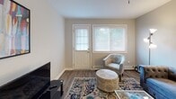 This single-floor residence offers a thoughtfully arranged layout that balances comfort and functionality throughout its key living spaces. The living room serves as the heart of the home, featuring warm hardwood-style flooring, soft gray walls, and multiple windows — including a glass-panel entry door — that allow natural light to fill the space. A tufted blue sofa, accent armchairs, and a colorful area rug contribute to a cozy yet stylish atmosphere.

Adjacent to the living area, the kitchen is equipped with gray shaker-style cabinetry, dark granite-look countertops, a mosaic tile backsplash, and a full suite of black appliances including a gas range, over-the-range microwave, and refrigerator. A double sink and a window overlooking greenery add to the kitchen's practicality. A dining table with vibrant upholstered chairs sits just off the kitchen, making the area ideal for everyday meals.

The two bedrooms are comfortable and well-lit, each featuring hardwood-style flooring, neutral walls, and windows with views of surrounding greenery. Both rooms are sized to accommodate a full bed with ease. A versatile third room with a window and wood flooring functions effectively as a home office or study. The full bathroom is clean and simply appointed, featuring a white vanity, toilet, and a shower-tub combination with a decorative curtain.