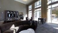 This stylish single-floor space exudes a modern, upscale ambiance defined by its dramatic soaring ceilings and expansive floor-to-ceiling windows that flood the interior with abundant natural light. The generous ceiling height creates an airy, open atmosphere that enhances the sense of spaciousness throughout.

The layout is thoughtfully organized into distinct functional zones. A plush, curved dark sectional sofa anchors the central lounge area, complemented by a sleek marble-topped coffee table and decorative accent pillows in navy and geometric patterns. Flowing sheer drapery softens the tall windows while maintaining the bright, welcoming atmosphere.

Toward the perimeter, intimate seating arrangements with upholstered chairs provide quieter conversation areas, while tall bar-height tables and chairs along the window line offer a more casual dining or workspace setting. Natural light pours through the glazing, offering views of the surrounding exterior.

A built-in shelving and display unit lines one wall, adding both visual interest and functional storage, while recessed and pendant lighting ensures the space is well-lit during evening hours. The neutral gray and charcoal color palette, combined with patterned carpeting and carefully curated furnishings, ties the entire space together into a cohesive, sophisticated aesthetic.