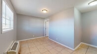 This single-floor unit presents a straightforward and functional layout finished with consistent light beige ceramic tile flooring throughout. The walls are painted in a neutral gray tone, complemented by crisp white baseboards that lend a clean, cohesive look to the interior.

The kitchen area is efficiently designed, featuring white cabinetry both above and below the counter, a double-basin stainless steel sink with a chrome faucet, granite-style countertops, and a white electric range. Upper cabinets provide ample storage, while the layout makes practical use of the available space.

The main living area is open and unfurnished, allowing for flexible use of the space. A white entry door with a side window fitted with horizontal blinds provides natural light and serves as the primary point of entry. A wall-mounted PTAC unit handles climate control, positioned beneath the window for efficient airflow throughout the room.

An interior door leads to an additional room, which is similarly finished with the same tile flooring, neutral gray walls, and a flush-mount ceiling light fixture. A six-panel interior door connects to what appears to be a utility or storage area, adding to the functional versatility of the unit. The overall ambiance is clean, minimal, and move-in ready.