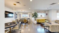 Located at 3483 SW 45th St in Gainesville, Florida, this single-floor community lounge is a thoughtfully designed social space that blends modern aesthetics with spirited University of Florida pride. The open layout is anchored by gleaming polished concrete floors that reflect natural light streaming in through multiple windows dressed with textured Roman shades, creating a bright and welcoming atmosphere throughout.

The space is divided into several distinct yet flowing zones. A comfortable lounge area features a bold chartreuse sofa, cream accent chairs, and a low coffee table, all arranged to encourage relaxed conversation. Nearby, a dedicated game area centers around a stylish round poker table beneath a contemporary multi-arm pendant light, accompanied by framed wall art and a flat-screen TV. A long communal dining table with modern upholstered chairs anchors a separate gathering zone, illuminated by a sleek brass-and-glass chandelier.

A full-service bar area is equipped with a rollup service window, dark countertops, bar seating with vibrant green woven stools, and a striking living moss wall installation as a dramatic backdrop. Lush potted indoor trees are positioned throughout, adding organic warmth to the contemporary interior.

Go Gators signage, vintage-style posters, and Gator-branded décor are woven seamlessly into the design, giving this lounge a lively, community-oriented character perfectly suited to its Gainesville location.