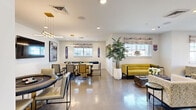 Located at 3483 SW 45th St in Gainesville, Florida, this single-floor community lounge is a thoughtfully designed social space that blends modern aesthetics with spirited University of Florida pride. The open layout is anchored by gleaming polished concrete floors that reflect natural light streaming in through multiple windows dressed with textured Roman shades, creating a bright and welcoming atmosphere throughout.

The space is divided into several distinct yet flowing zones. A comfortable lounge area features a bold chartreuse sofa, cream accent chairs, and a low coffee table, all arranged to encourage relaxed conversation. Nearby, a dedicated game area centers around a stylish round poker table beneath a contemporary multi-arm pendant light, accompanied by framed wall art and a flat-screen TV. A long communal dining table with modern upholstered chairs anchors a separate gathering zone, illuminated by a sleek brass-and-glass chandelier.

A full-service bar area is equipped with a rollup service window, dark countertops, bar seating with vibrant green woven stools, and a striking living moss wall installation as a dramatic backdrop. Lush potted indoor trees are positioned throughout, adding organic warmth to the contemporary interior.

Go Gators signage, vintage-style posters, and Gator-branded décor are woven seamlessly into the design, giving this lounge a lively, community-oriented character perfectly suited to its Gainesville location.
