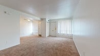 This two-floor unit offers a practical and straightforward layout designed for comfortable everyday living. The main level features a generously sized living area finished with warm beige carpeting throughout, white walls, and a ceiling fan with light fixture. Two large windows fitted with vertical blinds allow natural light to fill the space, while a structural beam adds subtle architectural definition between areas. The entry door is accessible directly from the living area. A carpeted staircase with a metal handrail leads to the upper floor, where additional living space continues the same neutral carpet and white-wall aesthetic. Bedrooms on the upper level are well-proportioned, with one featuring a sliding double-door closet and access to a bathroom. The bathroom is equipped with a single-sink vanity with white cabinetry, a tri-panel mirror medicine cabinet, tile flooring, and accessibility grab bars near the toilet and tub area. The kitchen is laid out in a galley configuration, featuring warm-toned wood cabinetry, dark laminate countertops, tile flooring, white appliances including a refrigerator and range, and a stacked washer/dryer unit conveniently integrated into the space.