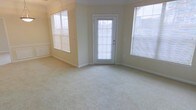This single-floor space features a spacious main living area designed with a light and airy aesthetic. The room is finished with wall-to-wall carpeting in a neutral cream tone, providing a soft underfoot feel that complements the overall warmth of the interior. The walls are painted in a soft yellow-beige hue, contributing to the inviting atmosphere throughout.

Natural light is a defining characteristic of this space, with multiple windows fitted with horizontal blinds positioned on both the left and right sides of the room, allowing for adjustable light control. A central glass-panel exterior door further enhances the brightness of the space while providing direct access to the outdoors.

Architectural detailing includes crown molding along the upper edges of the walls, adding a subtle but refined touch to the room's character. The clean white baseboards offer a crisp contrast against the carpeted floor. The layout of the room feels open and versatile, well-suited for a variety of furniture arrangements. Overall, the space presents a clean, move-in ready environment with a welcoming and functional design.