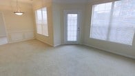 This single-floor space features a spacious main living area designed with a light and airy aesthetic. The room is finished with wall-to-wall carpeting in a neutral cream tone, providing a soft underfoot feel that complements the overall warmth of the interior. The walls are painted in a soft yellow-beige hue, contributing to the inviting atmosphere throughout.

Natural light is a defining characteristic of this space, with multiple windows fitted with horizontal blinds positioned on both the left and right sides of the room, allowing for adjustable light control. A central glass-panel exterior door further enhances the brightness of the space while providing direct access to the outdoors.

Architectural detailing includes crown molding along the upper edges of the walls, adding a subtle but refined touch to the room's character. The clean white baseboards offer a crisp contrast against the carpeted floor. The layout of the room feels open and versatile, well-suited for a variety of furniture arrangements. Overall, the space presents a clean, move-in ready environment with a welcoming and functional design.