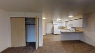 This single-floor unit features an open-concept layout that integrates the living area and kitchen into one cohesive space. The living area is finished with wall-to-wall carpet, providing a warm and comfortable feel underfoot, while the kitchen transitions to hard-surface flooring, offering a practical and easy-to-clean surface. The kitchen is well-equipped with white cabinetry providing ample storage, a built-in microwave, a refrigerator, and a stovetop. A peninsula extends from the cabinetry, creating additional counter space and a natural division between the kitchen and living area. Flush-mounted ceiling lights illuminate both spaces consistently throughout. A spacious closet is conveniently located within the living area, featuring a full hanging rod and generous depth for storage. A door visible toward the rear of the unit likely leads to additional rooms or an entry point. The overall aesthetic is clean and neutral, with white walls and light-toned finishes throughout, lending the space a bright and functional character well-suited for everyday living.