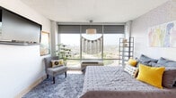 Located at 3900 Elati St in Denver, Colorado, this single-floor studio offers a thoughtfully arranged open layout that efficiently combines sleeping, living, dining, and working areas into one cohesive space. The sleeping area is a standout feature, anchored by large floor-to-ceiling windows that flood the room with natural light and frame expansive elevated views of the Denver cityscape and surrounding landscape. A plush area rug, a comfortable accent chair, and a decorative pendant light complete the relaxed yet stylish atmosphere of this zone. The living and dining area is defined by warm bamboo-style hardwood flooring, a wall-mounted TV, a compact media console, and a small dining table with chairs — providing functional spaces for both relaxation and everyday meals. A dedicated desk area offers a practical workspace, complemented by eclectic wall art that gives the studio a personalized, creative character. The bathroom is clean and well-appointed, featuring a large vanity with stone-look countertops, an oversized mirror, and a walk-in glass-enclosed shower with white tile surround. Throughout the studio, a mix of light and bold accent colors, layered textures, and unique decorative elements create a lively and inviting urban living environment.