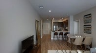This single-floor residence showcases a thoughtfully designed open-concept layout that seamlessly connects the living, dining, and kitchen areas into one cohesive, flowing space. Warm hardwood-style flooring runs throughout, anchoring the contemporary aesthetic and lending a sense of continuity from room to room.

The kitchen is a standout feature, equipped with dark cabinetry, stainless steel appliances including a full-sized refrigerator, and a light-colored countertop island that doubles as a breakfast bar. Three dark-toned bar stools line the island, creating a casual dining option. Recessed lighting is strategically placed overhead to illuminate the kitchen and adjoining areas with a clean, modern glow.

The living area offers ample room for comfortable furnishings, with a large flat-screen television serving as a focal point. A full-length mirror leans against the wall, enhancing the sense of openness and light in the space. The dining area, partially visible to the right, provides additional space for formal meals and entertaining.

Soft, neutral gray walls complement the warm flooring tones throughout, while the crisp white trim and interior doors add a clean architectural contrast. The overall ambiance is modern, functional, and inviting, making excellent use of the open floor plan to create a versatile living environment.