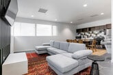 This thoughtfully designed single-floor space offers a seamless open-plan layout that integrates a living area, dining space, and fully equipped kitchen into one cohesive environment. The living area centers around a generous light grey sectional sofa anchored by a vibrant traditional area rug, creating a warm and inviting atmosphere. A striking dark slatted accent wall serves as a bold focal point, housing a wall-mounted flat-screen television that adds a contemporary media lounge feel. Recessed lighting throughout maintains a clean, bright ambiance, complemented by natural light filtering in through horizontal-blind-fitted windows along the perimeter walls. Large-format hexagonal tile flooring flows throughout the space, lending a modern and unified aesthetic. The kitchen is outfitted with rich dark wood cabinetry, open upper shelving, a stainless steel undermount sink, a gas range, and a full-size refrigerator. A distinctive hexagonal mosaic backsplash in grey and white tones serves as an eye-catching design detail. A dining table with wood chairs bridges the kitchen and living areas, making the space well-suited for group gatherings. The entry features glass-paneled doors with frosted detailing, offering a polished and welcoming first impression.