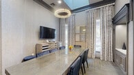 Located at 7009 Almeda Rd in Houston, TX, this sophisticated dining room delivers a refined and upscale atmosphere through thoughtful architectural detailing and high-end finishes. The centerpiece of the room is a large, polished stone-top dining table surrounded by navy blue upholstered chairs with a tailored grid pattern, offering seating for a full gathering. Overhead, a striking circular crystal chandelier anchors the space and adds a glamorous focal point beneath elevated ceilings accented with dark crown molding. The walls are dressed in a fine textured wallcovering in soft grey tones, lending the room a quiet elegance. A series of three built-in display niches with recessed lighting and raised-panel millwork line one wall, providing both architectural interest and display space. Floor-to-ceiling geometric-patterned drapes frame the windows, which allow generous natural light to filter in, offering views of an outdoor balcony. A wall-mounted flat-screen TV is positioned above a mirrored wood media credenza along one wall. The herringbone-patterned carpet with subtle green and gold accents adds warmth underfoot. A built-in sideboard with a white stone countertop completes the room's functional elegance, making this space ideal for formal dining and entertaining.