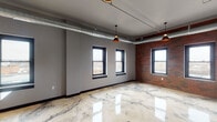 Located at 220 S Barstow St in downtown Eau Claire, Wisconsin, this single-floor unit blends industrial character with contemporary finishes throughout. The design aesthetic is defined by polished epoxy floors with striking marble-like veining that flows consistently across the entire space, reflecting light and creating a sense of expansiveness.

The open living and bedroom areas showcase exposed silver ductwork running along the ceilings, copper-toned pendant lighting, and black-framed windows that frame views of the surrounding cityscape. Original brick accent walls appear in the main living area, adding warmth and historic texture to the modern palette of cool gray walls.

The kitchen is well-appointed with dark espresso cabinetry, granite countertops, a large central island with a stainless steel sink, and a full suite of stainless steel appliances including a refrigerator, range, and microwave. Pendant lighting above the island enhances both functionality and style.

The bathrooms continue the space's cohesive design language, featuring granite-topped vanities with dark cabinetry, tub-shower combinations, and the same distinctive flooring. Throughout the unit, the interplay of industrial elements, natural materials, and refined finishes creates a polished yet characterful urban living environment.