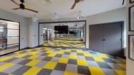 This dynamic fitness and movement studio is designed with both functionality and visual energy in mind. The most striking design element is the bold checkered carpet tile flooring, alternating between vivid yellow, gray, and neutral tones, creating a lively and motivating atmosphere throughout the open floor plan. Full-length mirrors span an entire wall, visually expanding the space and serving as a practical tool for movement and exercise form. A wall-mounted flat-screen television is positioned centrally along the mirrored wall, adding multimedia capability to the studio. The space is enclosed on one side by elegant steel-framed glass partition walls with matching black-framed pivot doors, offering visibility into an adjacent fitness equipment room while maintaining a sense of separation. Multiple ceiling fans are distributed across the room to support air circulation during active use, complemented by a series of pendant lights with polished metallic shades that provide focused illumination. Large windows along the perimeter bring in generous natural light and offer views of the surrounding urban environment. Framed artwork adds a thoughtful decorative touch to the windowed wall. The overall ambiance is energetic, modern, and well-appointed, making the studio well-suited for yoga, dance, group fitness classes, or personal training sessions.