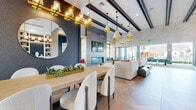 Located at 1575 Camino Altezza in Chula Vista, California, this single-story clubhouse is a thoughtfully designed community gathering space that blends contemporary aesthetics with warm, inviting materials. The architecture is defined by soaring exposed beam ceilings, stacked stone accent walls, and large-format herringbone-patterned tile flooring that unifies the entire space.

The main lounge area is expansive and light-filled, featuring floor-to-ceiling windows and folding glass walls that open fully to an outdoor terrace, creating a seamless indoor-outdoor living experience with views of palm trees and the surrounding community. Comfortable white sofas, leather ottomans, and eclectic pendant lighting in gold tones lend warmth and sophistication.

A dedicated dining area offers a substantial communal table surrounded by boucle-upholstered chairs beneath a striking gold chandelier, while a circular mirror anchors the adjacent dark-paneled accent wall. Nearby, an intimate lounge nook features rattan chairs, a black marble-topped bistro table, and a built-in banquette framed by teal cabinetry with illuminated open shelving.

The catering kitchen is well-appointed with warm wood cabinetry, white quartz countertops, under-counter refrigeration, and direct access to a bright secondary lounge area. A reception area near the entry provides a professional, welcoming arrival experience. Throughout, the design balances modern refinement with relaxed California living.
