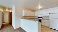 This single-floor residence offers a clean and functional layout designed for comfortable everyday living. The kitchen is a standout feature, showcasing an abundance of white cabinetry that provides generous storage space along both the upper and lower walls. A wraparound countertop in a neutral taupe tone extends into a peninsula, creating a natural divide between the kitchen and the adjacent living area while also offering additional prep and dining surface. The kitchen is equipped with a white electric range and an under-cabinet range hood, complementing the bright, cohesive aesthetic. Tile flooring in the kitchen transitions to carpet in the living space, adding warmth and definition between the two areas. The overall color palette is light and neutral — white walls, soft beige tones, and warm lighting from a flush-mount ceiling fixture — contributing to a bright and welcoming ambiance throughout. The open-concept flow between the kitchen and living space makes the home feel spacious and well-connected, ideal for both everyday routines and casual entertaining. The space presents a straightforward, move-in ready environment with a timeless and practical design sensibility.