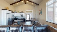This single-floor community space is designed for comfort and versatility, making it an ideal gathering area for socializing and events. The room's most striking architectural feature is its soaring vaulted ceiling accented by rich, dark exposed wood beams, which lend a warm, rustic-modern character to the space. Recessed lighting is thoughtfully positioned throughout to ensure the interior remains bright and welcoming at any hour.

The layout accommodates a variety of uses, with a lounge area anchored by a substantial leather sofa, complemented by multiple round bistro-style tables paired with industrial stools — offering both casual seating and dining options. Dark hardwood flooring runs throughout, grounding the open layout with a sense of warmth and continuity.

Along one wall, a well-appointed kitchenette features white cabinetry, dark countertops, a sink, and full-size appliances, providing all the essentials for hosting and refreshments. Natural light pours into the space through a set of elegant French doors at the far end, which connect the interior to the outdoors and enhance the airy, open ambiance. The neutral wall tones and tasteful decorative accents complete the polished yet approachable aesthetic of this functional communal space.