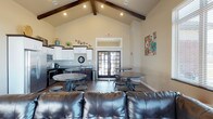This single-floor community space is designed for comfort and versatility, making it an ideal gathering area for socializing and events. The room's most striking architectural feature is its soaring vaulted ceiling accented by rich, dark exposed wood beams, which lend a warm, rustic-modern character to the space. Recessed lighting is thoughtfully positioned throughout to ensure the interior remains bright and welcoming at any hour.

The layout accommodates a variety of uses, with a lounge area anchored by a substantial leather sofa, complemented by multiple round bistro-style tables paired with industrial stools — offering both casual seating and dining options. Dark hardwood flooring runs throughout, grounding the open layout with a sense of warmth and continuity.

Along one wall, a well-appointed kitchenette features white cabinetry, dark countertops, a sink, and full-size appliances, providing all the essentials for hosting and refreshments. Natural light pours into the space through a set of elegant French doors at the far end, which connect the interior to the outdoors and enhance the airy, open ambiance. The neutral wall tones and tasteful decorative accents complete the polished yet approachable aesthetic of this functional communal space.