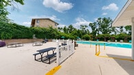 This outdoor recreational facility centers around a generously sized swimming pool with crystal-clear turquoise water, framed by a wide concrete deck that provides ample space for lounging. The deck is furnished with teal pool chairs and shaded by colorful umbrellas, creating a comfortable environment for sunbathers and swimmers alike. A decorative wooden pergola sits at one end of the pool area, offering additional shaded seating. The facility is enclosed by a white vinyl fence with a gated entry, accented by raised stone planters with ornamental grasses that add a polished landscaping touch. A single-story brick amenity building runs along one side of the pool and features a covered walkway with support columns, providing shade and shelter. This building houses restrooms, trash receptacles, and a vending machine for convenience. Picnic tables are positioned throughout the deck area for casual outdoor dining. The surrounding landscape is lush with mature trees that provide a natural green backdrop, enhancing the sense of privacy and seclusion. A multi-story brick residential building is visible beyond the tree line, indicating that this pool complex serves as a shared amenity. The overall atmosphere is inviting and well-maintained, designed to support active outdoor leisure.