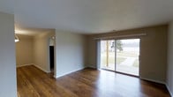 This single-story space presents a clean, straightforward layout centered around a bright and open living area. The room is anchored by rich, dark hardwood flooring that extends throughout, adding warmth and visual depth to the otherwise neutral interior. Walls are finished in a soft, light tone, complemented by white baseboards that provide a crisp contrast.

A prominent sliding glass door dominates one wall of the living area, serving as both a functional entry point and a significant source of natural light. The door opens to a view of an outdoor patio and yard, with mature trees and greenery visible beyond, lending a sense of connection to the exterior environment. Sheer white curtains frame the glass door, softening the transition between indoors and outdoors.

The ceiling features a subtle textured finish and includes a visible ventilation register, indicating a ducted HVAC system. A doorway visible on the adjacent wall leads to a separate area of the home, suggesting a practical and connected floor plan. The space is unfurnished, allowing the architectural elements and natural light to define the overall ambiance, which feels calm, open, and functional.