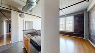 Located on East Broad Street in Richmond, VA, this one-bedroom, two-bathroom loft showcases a compelling blend of industrial character and modern comfort. The open-concept living area is anchored by warm hardwood floors, soaring ceilings with exposed ductwork and structural beams, and striking original brick walls that add texture and historic charm throughout. Large windows fitted with horizontal blinds flood the space with natural light while framing views of the surrounding urban streetscape.

The kitchen features granite countertops, warm wood cabinetry, stainless steel appliances including a refrigerator, microwave, and range, and a peninsula with a double sink and dishwasher — all set beneath dramatic pendant lighting and oversized HVAC ductwork that reinforces the loft's industrial aesthetic.

The bedroom continues the hardwood flooring and exposed brick theme, with generous windows and access to a closet with built-in shelving. Two bathrooms are finished with slate-look tile flooring, walk-in tiled showers, and clean white fixtures. Additional closet space and multiple hallways provide practical storage and circulation throughout.

Ceiling fans in the living and bedroom areas add comfort, while the overall design strikes a balance between raw, warehouse-inspired architecture and thoughtfully modern finishes.