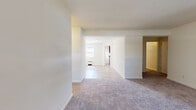 This single-floor unit offers a clean, functional layout designed for comfortable everyday living. The main living area features soft wall-to-wall carpeting and flows seamlessly into an adjoining room through a wide, open passageway, creating a connected and airy feel throughout. Multiple windows dressed with blinds allow natural light to filter in across the living and bedroom spaces, each of which is equipped with baseboard or wall-mounted radiators for year-round comfort.

The kitchen is a standout feature, outfitted with rich, dark wood cabinetry, granite-style countertops, a stainless steel sink, and a full-size refrigerator. Light wood-style flooring runs through the kitchen, lending warmth and easy maintenance to the space. A window above the sink brings in additional natural light and overlooks the surrounding greenery.

The bedrooms are generously proportioned, each with plush carpeting and large windows that frame views of mature trees outside. The bathroom is finished with classic white subway tile wainscoting, wood-look flooring, a soaking tub, and a frosted glass window for privacy while still admitting natural light. Throughout the unit, neutral cream-toned walls provide a versatile backdrop ready for personalization.