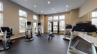This single-story fitness center is thoughtfully designed to accommodate a full range of workout routines across multiple interconnected areas. The main exercise space features soaring ceilings and large multi-pane windows that flood the room with natural light, creating an open and energizing atmosphere. Warm neutral-toned walls complement the durable dark rubber flooring found throughout the facility.

The primary cardio area is well-stocked with Matrix treadmills, elliptical trainers, and a Precor recumbent bike, alongside a cable resistance machine and adjustable weight bench. A wall-mounted flat-screen television adds convenience for those looking to stay entertained during workouts. A large mirror runs along one wall, visually expanding the space and providing form-checking functionality.

A separate strength and flexibility room, finished in a cool blue-gray palette, offers a more focused training environment. This area includes an adjustable weight bench, a rack of dumbbells in varying weights, medicine balls, balance trainers, aerobic step platforms, and a large wall mirror. Tall windows provide natural light while maintaining privacy.

Together, the two areas deliver a comprehensive and well-organized fitness environment suited for cardiovascular training, strength work, and functional fitness.