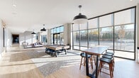 Located at 417 Callowhill Street in Philadelphia's vibrant Callowhill neighborhood, this expansive single-floor amenity lounge offers a generously sized, open-plan layout designed for social interaction and relaxation. The space is defined by dramatic floor-to-ceiling curtain-wall glazing that spans the full perimeter, flooding the interior with abundant natural light and framing street-level views of the surrounding neighborhood.

Light hardwood flooring runs throughout, complementing the neutral, contemporary palette of white and warm gray tones on the walls and ceilings. Soaring ceiling heights enhance the airy, loft-like atmosphere. The lounge area features plush, modular sectional sofas in muted mauve and taupe tones, arranged atop geometric patterned area rugs alongside solid wood coffee tables, creating inviting conversation zones bathed in sunlight.

A dedicated recreation area houses a full-size billiards table centered on its own rug, accompanied by bar-height communal tables and seating. Oversized black dome pendant lights punctuate the ceiling, providing focused illumination that complements the recessed lighting throughout. A kitchenette or bar area with dark countertops and white cabinetry offers additional functionality. The space connects seamlessly to an outdoor area through large glass entry doors, reinforcing the indoor-outdoor connection that defines this modern, community-focused amenity space.
