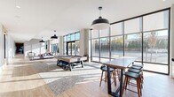 Located at 417 Callowhill Street in Philadelphia's vibrant Callowhill neighborhood, this expansive single-floor amenity lounge offers a generously sized, open-plan layout designed for social interaction and relaxation. The space is defined by dramatic floor-to-ceiling curtain-wall glazing that spans the full perimeter, flooding the interior with abundant natural light and framing street-level views of the surrounding neighborhood.

Light hardwood flooring runs throughout, complementing the neutral, contemporary palette of white and warm gray tones on the walls and ceilings. Soaring ceiling heights enhance the airy, loft-like atmosphere. The lounge area features plush, modular sectional sofas in muted mauve and taupe tones, arranged atop geometric patterned area rugs alongside solid wood coffee tables, creating inviting conversation zones bathed in sunlight.

A dedicated recreation area houses a full-size billiards table centered on its own rug, accompanied by bar-height communal tables and seating. Oversized black dome pendant lights punctuate the ceiling, providing focused illumination that complements the recessed lighting throughout. A kitchenette or bar area with dark countertops and white cabinetry offers additional functionality. The space connects seamlessly to an outdoor area through large glass entry doors, reinforcing the indoor-outdoor connection that defines this modern, community-focused amenity space.