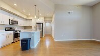 This thoughtfully designed 1-bedroom, 1-bathroom unit offers a comfortable and functional single-floor layout. The open-concept living and kitchen area is anchored by a central island with pendant lighting overhead, complemented by crisp white cabinetry, stainless steel appliances — including a refrigerator, range, and microwave — and a clean, modern aesthetic throughout. Hardwood-style flooring runs through the main living spaces, reflecting natural light and adding warmth to the interior. The living area is bright and open, featuring a ceiling fan, sliding glass doors, and a transom window above that floods the space with natural light and provides access to a private balcony with pleasant outdoor views. A hallway with built-in storage connects the main living area to the private quarters. The bedroom offers plush carpeting, a large window with blinds, and a sliding-door closet with double hanging rods for ample storage. The full bathroom is finished with a white vanity, tile-surround tub and shower combination, and hardwood-style flooring, with a small window allowing in natural light. The unit's neutral color palette and clean finishes create a bright, move-in-ready environment throughout.
