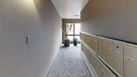 This mail area is a well-organized, single-floor corridor designed to serve the residents of the building efficiently. The hallway is lined along one side with a full bank of individual tenant mailboxes finished in a classic gold tone, providing convenient mail access for all units. The flooring throughout the corridor is terrazzo-style, offering a durable and clean surface that complements the neutral, taupe-toned walls.

Natural light filters in through large windows at the end of the hallway, brightening the space and offering views of the surrounding exterior grounds, including landscaping and adjacent structures. The windows are framed in dark trim, adding a subtle contrast to the light-colored walls. Decorative potted plants — including tall, sculptural varieties — are positioned near the windows, introducing a touch of greenery and warmth to the utilitarian space.

Overhead, flush-mounted ceiling lights and wall-mounted sconces provide consistent illumination throughout the corridor. An exit sign marks the pathway, and unit doors — such as the teal-painted door to unit 102 — are visible along the route, reinforcing the building's residential character. The overall ambiance is clean, practical, and thoughtfully maintained, creating a pleasant everyday environment for residents collecting their mail.