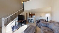 Located in Pewaukee, Wisconsin, this two-story space showcases an open, airy design characterized by soaring vaulted ceilings and an abundance of natural light. The main level centers around a welcoming living area with plush carpeting, where a staircase with metal balusters leads to the upper floor. Large windows and an elevated clerestory window draw in generous daylight, while a ceiling fan enhances comfort throughout. The living area flows seamlessly toward a kitchen featuring warm wood cabinetry, light countertops, tile flooring, and a breakfast bar with seating — ideal for casual dining.

Ascending the staircase, the upper level reveals a spacious loft-style primary bedroom with vaulted ceilings, a ceiling fan, and a connected en-suite bathroom. The bathroom is finished with tile flooring, a wood vanity, and a window that provides natural light. An additional bedroom on the upper level offers carpeted floors, a bright window with blinds, and a calm, neutral palette. Throughout the space, a consistent use of soft neutral tones, carpet, and tile creates a cohesive and comfortable atmosphere well-suited for everyday living.