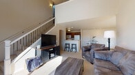 Located in Pewaukee, Wisconsin, this two-story space showcases an open, airy design characterized by soaring vaulted ceilings and an abundance of natural light. The main level centers around a welcoming living area with plush carpeting, where a staircase with metal balusters leads to the upper floor. Large windows and an elevated clerestory window draw in generous daylight, while a ceiling fan enhances comfort throughout. The living area flows seamlessly toward a kitchen featuring warm wood cabinetry, light countertops, tile flooring, and a breakfast bar with seating — ideal for casual dining.

Ascending the staircase, the upper level reveals a spacious loft-style primary bedroom with vaulted ceilings, a ceiling fan, and a connected en-suite bathroom. The bathroom is finished with tile flooring, a wood vanity, and a window that provides natural light. An additional bedroom on the upper level offers carpeted floors, a bright window with blinds, and a calm, neutral palette. Throughout the space, a consistent use of soft neutral tones, carpet, and tile creates a cohesive and comfortable atmosphere well-suited for everyday living.