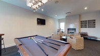 Located at 5927 Avery Wood Ave in Dublin, Ohio, this single-story community clubhouse offers a well-appointed gathering space designed for both leisure and social connection. The main area seamlessly blends entertainment and comfort, anchored by a full-size billiards table positioned beneath a striking geometric chandelier that adds a modern, refined touch to the room. A wall-mounted flat-screen television provides additional entertainment options for residents and guests. Toward the back of the space, a cozy lounge area invites relaxation, featuring plush neutral-toned armchairs and a coffee table arranged around a sleek fireplace with a tiled surround, creating a warm and inviting atmosphere. Natural light filters in through a window adjacent to the fireplace, brightening the space and enhancing its open, airy feel. The interior is finished with clean white walls, recessed lighting, and a combination of hard and carpeted flooring that delineates activity zones. A high-top table near the window offers an additional spot for casual conversation. The overall design balances functionality with sophistication, making this clubhouse an ideal amenity for everyday use and community gatherings alike.