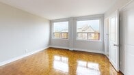 This efficient one-bedroom, one-bathroom unit makes smart use of its intimate footprint across a single floor. The living and sleeping areas showcase warm, honey-toned parquet flooring in a classic block pattern, polished to a high sheen that reflects natural light throughout the space. Large windows in both the main living area and bedroom invite generous daylight and frame views of the surrounding neighborhood streetscape and architectural details beyond.

The kitchen is thoughtfully appointed with rich, dark-stained wood cabinetry extending to the ceiling for maximized storage. Stainless steel appliances — including a full-sized refrigerator, electric range, and integrated range hood — sit against a subway tile backsplash, while slate-toned floor tiles add contrast and durability underfoot. A single-basin sink with modern fittings completes the functional layout.

The bedroom offers a clean, neutral palette with bright white walls and the same gleaming parquet flooring, creating a calm and restful atmosphere. The bathroom is finished with light beige ceramic floor tiles, a pedestal sink with chrome fixtures, a toilet, and a tiled shower-tub combination. A frosted window provides natural ventilation while maintaining privacy. Clean white trim and simple, classic detailing carry throughout the entire unit, lending a cohesive and well-maintained character to the space.
