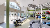 This comprehensive fitness center is thoughtfully designed across a single floor, offering a wide range of dedicated workout zones to accommodate various exercise disciplines. The facility features soaring vaulted ceilings in the main training area, creating an open and energizing atmosphere, while large floor-to-ceiling windows and glass doors flood the space with natural light and provide direct views of the adjacent outdoor pool and patio.

The cardio zone houses a row of modern treadmills and elliptical machines, complemented by a motivational feature wall displaying inspirational quotes. A functional training area is anchored by a TuffStuff Fitness overhead rig, a Ringside heavy punching bag, and a WorX rack system stocked with medicine balls, kettlebells, and TRX suspension trainers set against vibrant green accent walls. Additional equipment includes strength machines, a power tower, mini trampolines, and a tire sled for conditioning work.

Full-length mirrors are positioned throughout to support proper form, and ceiling fans ensure comfortable airflow. The flooring transitions between carpet tile and rubberized surfaces across zones. Glass doors open directly to a sunlit pool deck, seamlessly connecting indoor training with outdoor recreation. The facility's bold green and white color palette reinforces an energetic, performance-focused environment throughout.