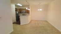 This single-floor space is designed with an open-concept layout that seamlessly connects the kitchen, dining, and living areas, creating a cohesive and functional living environment. The kitchen features warm-toned wooden cabinetry, granite countertops, and stainless steel appliances, including a refrigerator and over-the-range microwave. A peninsula with a raised breakfast bar serves as both a practical workspace and a natural divider between the kitchen and the adjacent living and dining spaces. The kitchen area is illuminated by recessed fluorescent lighting and a trio of modern pendant lights suspended over the peninsula, adding a stylish focal point. The transition into the dining and living areas is marked by a shift from hardwood-style flooring to plush, neutral-toned carpeting, which adds warmth and comfort underfoot. An elegant multi-arm chandelier anchors the dining area, providing a refined touch to the space. The walls are finished in a soft, neutral tone throughout, complementing the overall palette. Crown molding accents the ceilings, lending the interior a polished, well-appointed feel across the entire open floor plan.