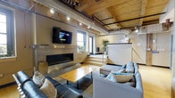 This single-floor space showcases a compelling industrial-chic aesthetic, blending raw architectural elements with refined modern finishes. The living area draws immediate attention with its exposed metal ductwork and warm wooden ceiling beams, creating a loft-like atmosphere that balances rugged character with contemporary comfort. Track lighting runs along the ceiling, providing both functional illumination and an added layer of visual interest.

A sleek, horizontal fireplace is mounted low on the wall, framed by a minimalist surround and complemented by a wall-mounted flat-screen television above. The seating area features a generous sectional sofa in neutral grey tones accented with colorful throw pillows, arranged around a natural wood-topped coffee table that adds organic warmth to the space.

A raised platform area at the rear of the room, accessible via a short set of stairs, introduces a subtle sense of spatial layering. Natural light filters in through white-framed windows, enhancing the open, airy feel. Potted greenery adds a welcoming, natural touch throughout. The overall ambiance is modern, relaxed, and thoughtfully designed for everyday living.