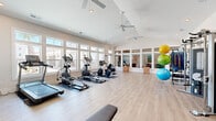 This fitness center offers a bright, airy environment designed to support a full range of workout activities. The space is defined by its striking vaulted ceilings, which lend a sense of openness and volume, complemented by multiple ceiling fans that ensure comfortable air circulation throughout. Large, multi-pane windows line the walls, flooding the interior with natural light and providing views of the surrounding exterior, creating an inviting atmosphere for exercise.

The light wood-tone flooring runs throughout, contributing to the clean, modern aesthetic of the space. Cardio equipment is well-represented, with treadmills, elliptical trainers, stationary bikes, and a cycling bike arranged along the windowed walls. A dedicated strength training area features a multi-function cable machine, a dumbbell rack with a full range of weights, and an adjustable workout bench. Stability balls in multiple sizes are neatly stored on a rack, offering options for flexibility and core training.

A wall-mounted flat-screen television is positioned for visibility across the fitness floor. A mirrored section along one wall enhances the sense of space and supports form-checking during workouts. The overall design balances functionality with a welcoming, light-filled ambiance, making it a well-rounded facility for both cardio and strength-focused training.