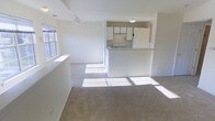 This two-floor residence offers a clean, neutral aesthetic with a welcoming open-concept layout on the main level. The living area is generously proportioned, finished with plush wall-to-wall carpeting in a warm beige tone that adds comfort underfoot. Large windows allow abundant natural light to pour in, casting bright, airy patches across the floor and enhancing the sense of openness throughout the space.

The kitchen is positioned at the heart of the main level, separated from the living area by a raised breakfast bar that serves as both a functional workspace and a casual dining surface. White cabinetry lines the kitchen walls, providing ample storage, while the white appliances and under-cabinet range hood contribute to a cohesive, bright finish. The pass-through design of the kitchen maintains a visual connection to the living space, making the layout feel connected and sociable.

Recessed track lighting on the ceiling offers supplemental illumination, complementing the natural light from the windows. The crisp white walls throughout create a fresh, move-in-ready canvas. With two floors, this residence provides well-defined areas for living and private functions, making it a practical and comfortable home.