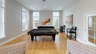 This single-floor social space is designed for recreation and relaxation, offering a well-appointed game room environment. The layout flows between distinct activity zones, each thoughtfully furnished to encourage gathering and entertainment.

The main game area showcases hardwood flooring and is anchored by a full-size billiards table with pendant lighting overhead, while a shuffleboard table lines one wall, providing additional gaming options. An upright arcade cabinet adds a nostalgic touch to the space. Large double-hung windows throughout flood the interior with natural light and offer views of the surrounding greenery outdoors.

A comfortable lounge area transitions from the game zone, featuring patterned upholstered chairs and a carpet area rug that define the seating space. Warm accent walls in golden yellow contrast with the neutral gray tones found throughout the rest of the room, creating visual interest. Abstract artwork decorates the walls, contributing to the contemporary aesthetic.

A French door entry connects the space to adjacent areas, with high ceilings adding to the open, airy atmosphere. Recessed lighting supplements the natural light, ensuring the space remains well-lit throughout the day and evening.