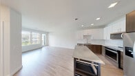 This contemporary single-floor unit offers a bright and open layout designed with clean lines and modern finishes. The open-concept living and kitchen area is the heart of the space, featuring light hardwood-style flooring that flows seamlessly throughout. Large windows and glass doors invite abundant natural light, with views of the surrounding neighborhood enhancing the airy atmosphere.

The kitchen is well-appointed with a two-tone cabinetry scheme — white upper cabinets contrast elegantly with rich dark wood lower cabinets — set against a white tile backsplash. Stainless steel appliances include a refrigerator with ice maker, a range with oven, a built-in microwave, and a dishwasher. The stainless steel undermount sink is paired with a sleek industrial-style faucet. A central island with a stainless steel countertop and integrated wine rack adds both functionality and visual interest.

The bedroom is generously sized, bathed in natural light from a large picture window, and features the same warm hardwood-style flooring. A closet provides practical storage. The bathroom maintains the modern aesthetic with a floating dark wood vanity, white countertop, and large mirror, complemented by neutral tile flooring. Recessed lighting throughout ensures a well-lit, polished finish across the entire unit.