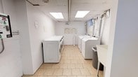 This shared laundry facility offers a practical and well-maintained space designed to accommodate multiple users efficiently. The room features a lineup of both front-loading and top-loading washing machines along one wall, complemented by a matching set of dryers, providing ample capacity for laundry needs. The space is finished with light-toned tile flooring that is easy to clean and maintain, contributing to the overall bright and tidy atmosphere. Overhead, a drop ceiling with recessed fluorescent panel lighting ensures the room is evenly and thoroughly illuminated. The walls are painted white, reinforcing the clean and functional aesthetic throughout. A utility sink is positioned conveniently in the corner, offering additional practicality for users. Ventilation ductwork is visible along the upper wall near the dryers, ensuring proper airflow and safe operation of the machines. Posted instructions and notices on the wall keep users informed of proper usage guidelines. A waste bin is also provided for convenience. The layout is thoughtfully arranged to allow comfortable movement between machines, making the laundry process straightforward and efficient.