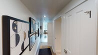 This single-floor urban residence offers a thoughtfully designed layout that balances modern style with everyday functionality. The entryway greets visitors with a well-lit corridor adorned with framed artwork, hardwood-style flooring, and a runner rug, establishing an inviting transition into the living spaces beyond.

The kitchen is a standout feature, showcasing white shaker cabinetry, granite countertops, a subway tile backsplash, and a full suite of stainless steel appliances including a refrigerator, range, microwave, and dishwasher. A central island with a marble-style surface provides additional workspace and casual dining space.

The living area is bathed in natural light through floor-to-ceiling windows that frame sweeping high-rise city views, creating a dramatic urban backdrop. Wood-style flooring runs throughout, complementing the contemporary furnishings and open feel of the space.

The bathroom is finished in a clean, neutral palette with large-format tile flooring, white cabinetry, granite countertops, a well-lit vanity mirror, and a shower-tub combination — all contributing to a calm, spa-like atmosphere.

The bedroom, visible in the dollhouse view, features a warm, layered design with open shelving and ample natural light. Overall, the residence presents a cohesive, modern aesthetic ideal for urban living.
