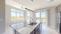 This contemporary single-floor unit is thoughtfully designed with a clean, neutral palette and light-toned luxury vinyl plank flooring throughout. The open-concept kitchen serves as the heart of the space, showcasing dark cabinetry with brushed nickel hardware, sweeping granite countertops, a white subway tile backsplash, and a full suite of stainless steel appliances including a range, microwave, refrigerator, and dishwasher. A central kitchen island with an integrated sink and pendant lighting above adds both style and functionality. Large windows and a sliding glass door in the kitchen and living area flood the space with natural light and provide direct access to a private balcony, which overlooks open green landscapes. The bedrooms are well-proportioned, featuring large windows—some floor-to-ceiling—that bring in abundant sunlight and offer views of the surrounding area. Each bedroom includes closet storage, and at least one features a private en-suite bathroom and dedicated closet space with built-in shelving. Ceiling fans provide comfort in key living areas. The overall aesthetic is modern and move-in ready, with a cohesive design language that emphasizes light, openness, and practical living.