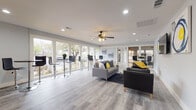 Located at McCain Park Dr in North Little Rock, Arkansas, this single-floor community space blends modern design with practical functionality. The open-concept layout is anchored by a spacious lounge area finished with light gray luxury vinyl plank flooring throughout, crisp white walls, and recessed lighting complemented by a ceiling fan with a light fixture. The lounge is furnished with a gray sectional sofa, leather accent chairs, and a wall-mounted flat-screen TV, all accented by a bold blue and yellow color palette reflected in the artwork and décor. A row of bar-height tables and stools lines the large windows along one wall, flooding the room with natural light and offering views of the surrounding greenery. The fully equipped kitchen features white shaker-style cabinetry, dark countertops, a classic subway tile backsplash, and a complete suite of stainless steel appliances including a refrigerator, range, microwave, and dishwasher. Yellow accent rugs add a vibrant touch to the kitchen floor. A fitness area is also present within the space, housing exercise equipment for residents' use. The overall ambiance is clean, bright, and inviting, making this a well-appointed communal facility designed to accommodate both relaxation and active use.