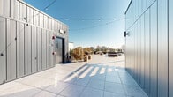 This expansive rooftop terrace offers a well-designed outdoor amenity space with striking panoramic views of the surrounding city skyline. The terrace is finished throughout with large-format stone-look tiles that lend a clean, contemporary aesthetic underfoot, while slatted wood privacy screens and modern metal panel structures define the perimeter and entry points.

The space is organized into several distinct zones. A casual lounge area features stylish woven egg chairs paired with concrete side tables, ideal for relaxed seating. String lights are strung overhead, adding a warm, inviting ambiance for evening use. A dedicated dining area sits beneath a bold dark-framed pergola, which provides partial shade while maintaining an open-air feel, and is equipped with an outdoor television mounted to the wall — perfect for entertaining.

Additional seating is scattered throughout the terrace, including low-profile sofas and modular lounge furniture arranged to take advantage of the unobstructed skyline views. Potted greenery and planters line the railings, softening the architectural edges with natural elements.

The terrace benefits from abundant natural light and open sky exposure, making it a versatile space suited for social gatherings, casual lounging, or al fresco dining throughout the day and into the evening.