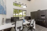 This single-floor business center is thoughtfully designed to offer a functional and contemporary workspace environment. The room features a continuous white L-shaped desk surface accommodating multiple workstations, each equipped with desktop computers and ergonomic mesh office chairs, making it well-suited for individual or shared use. Dark tile flooring with a large-format grid pattern runs throughout the space, providing a sleek and durable foundation that complements the neutral white walls. Overhead, a cluster of modern pendant-style spotlights adds focused ambient illumination, while a dedicated recessed downlight serves additional areas of the room. Large floor-to-ceiling windows along one wall allow generous natural light to pour in, offering views of a lush outdoor greenscape that enhances the overall brightness and openness of the space. Built-in cabinetry with a gray wood-tone finish provides convenient storage alongside a dedicated printer station. A standout feature is the Luxer One smart package locker system — a tall, multi-compartment black unit that offers secure, automated package management. Abstract blue and teal canvas artwork adorns the walls, adding a refined artistic touch. A frosted-glass door provides privacy while maintaining a sense of openness, completing this well-appointed, modern space.