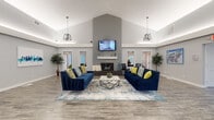 Located at 4365 Bayshire Rd in Groveport, Ohio, this single-floor community clubhouse offers a thoughtfully designed gathering space that balances comfort, functionality, and modern style. The centerpiece of the space is a spacious open lounge area with soaring vaulted ceilings that create an airy, expansive atmosphere. Two deep navy blue sofas, accented with yellow and patterned throw pillows, are arranged symmetrically around a geometric marble-top coffee table and a soft area rug, establishing an inviting and polished social hub. A floor-to-ceiling gray accent wall frames a sleek linear fireplace with a white mantel, above which a wall-mounted flat-screen television is positioned. Industrial-style pendant lights hang throughout, adding a contemporary touch. Adjacent to the lounge, a dedicated workstation area features multiple desks and ergonomic chairs, providing a productive space for residents. A kitchenette equipped with stainless steel appliances, a counter-height island, and bar seating offers convenient amenities for gatherings and day-to-day use. The entire space features durable wood-look plank flooring in a neutral gray tone, complemented by large windows dressed with white blinds that allow natural light to flow throughout. Double glass entry doors with a branded welcome mat complete the welcoming, well-appointed interior.