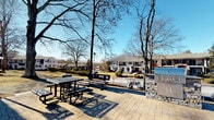 The Fire Pit area is a well-appointed outdoor amenity space designed for community gathering and relaxation. The foundation of this space is a generously sized stone-tile patio, laid in a neutral palette that complements the natural surroundings. The area is framed by the building's striking exterior, characterized by bold terracotta-orange architectural panels, large floor-to-ceiling windows, and clean white trim — creating a modern yet welcoming aesthetic.

The outdoor space is thoughtfully furnished with metal picnic tables and benches, providing ample seating for casual dining or socializing. A stone-clad built-in grill station serves as a functional centerpiece, ideal for outdoor cooking. Curved pathways extend across the patio, connecting to additional outdoor lounge seating areas outfitted with cushioned sofas and chairs, perfect for relaxed conversation.

Beyond the patio, the landscape opens to views of mature trees, manicured lawns, and neighboring residential buildings, offering a tranquil, park-like backdrop. White market umbrellas near what appears to be a pool area are visible in the distance, suggesting a broader outdoor amenity complex. The space benefits from abundant natural light and open skies, making it an ideal setting for outdoor gatherings throughout the seasons. Large glass entry doors provide seamless indoor-outdoor access directly from the building.